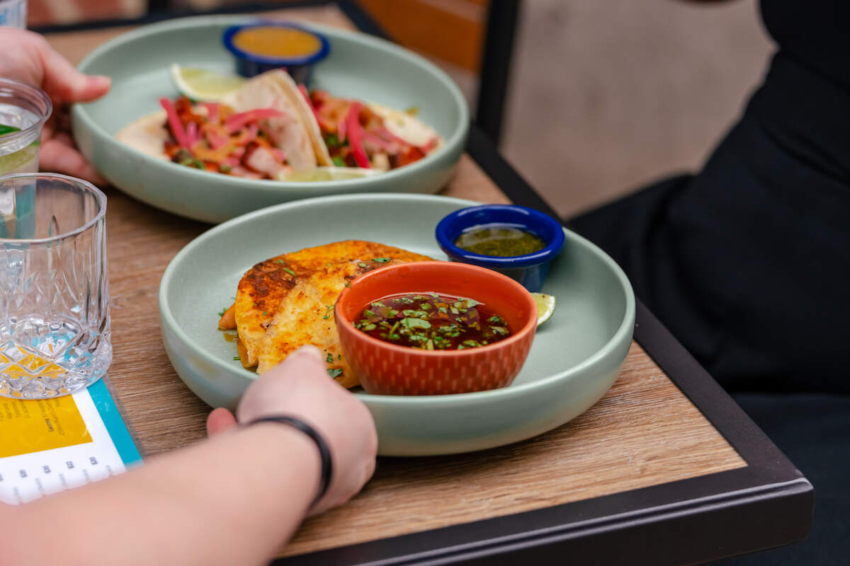 A close-up shot features two light-green plates of Mexican food served on a wooden table, with a person’s hand reaching towards the foreground dish. The front plate holds crispy, golden-brown quesadillas or birria tacos, accompanied by a small blue bowl of green salsa and a larger orange bowl filled with dipping broth topped with fresh cilantro. In the blurred background, another plate showcases tacos generously garnished with pickled red onions and fresh herbs.