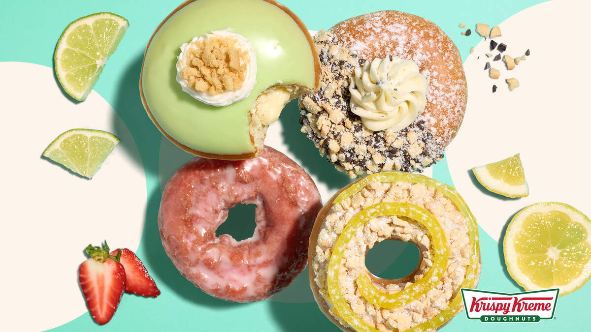 A top-down view features four vibrant Krispy Kreme doughnuts arranged on a bright teal and cream geometric background. The selection includes a pink glazed doughnut, a lemon-yellow ring with a icing swirl, a sugar-dusted doughnut with cream and cookie crumbles, and a light green iced doughnut with a bite taken out to reveal a creamy filling. Scattered fresh strawberry slices, lime wedges, lemon wheels, and cookie crumbs accent the corners, with the Krispy Kreme logo positioned in the bottom right.