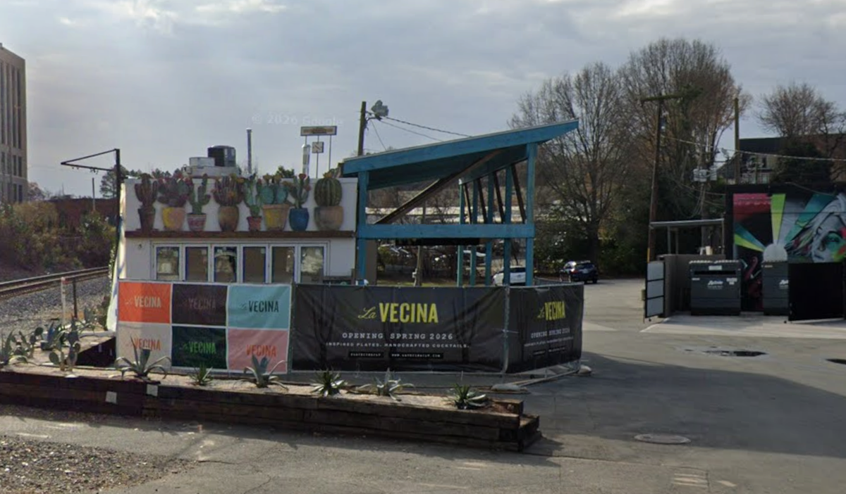 A street-level view shows a modern restaurant called La Vecina, featuring a black banner that announces its opening in spring 2026 alongside colorful murals of cacti and abstract figures. The building is situated next to a railroad crossing signal and a small planter filled with agave plants. In the background, a multi-story apartment complex sits under a cloudy sky, completing the urban scene.