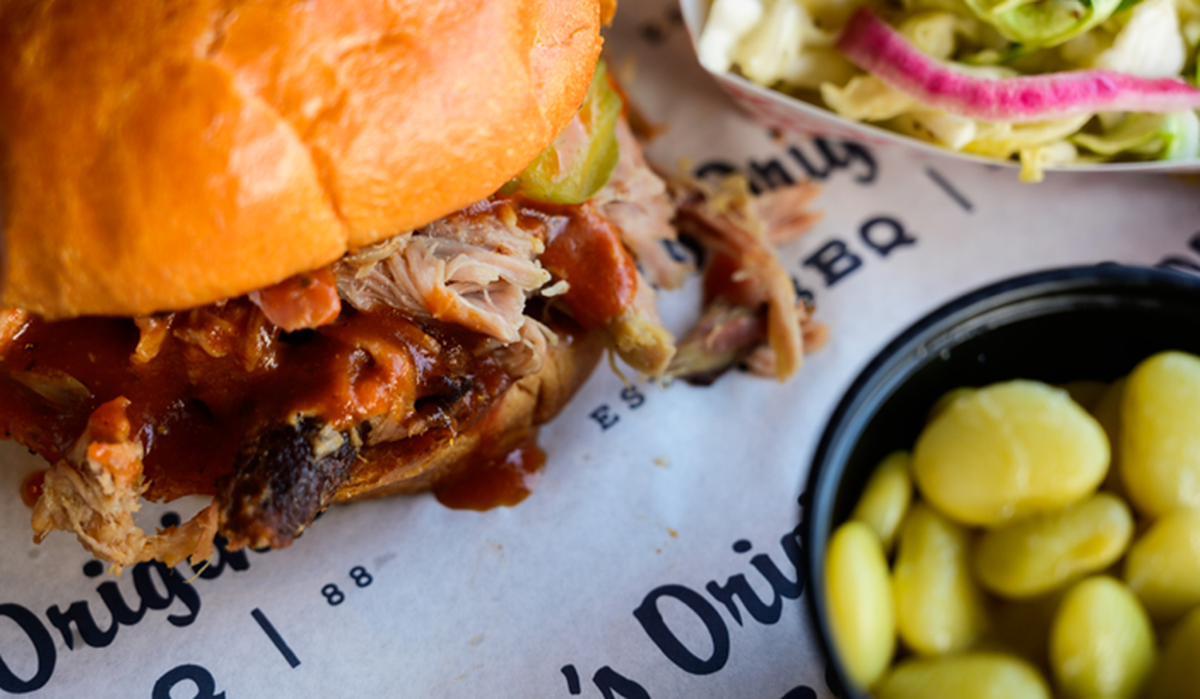 A towering pulled pork sandwich is served on a glossy toasted bun, with juicy shreds of meat dripping in a rich, dark barbecue sauce. In the soft-focus background, a side of creamy coleslaw with red onions and a small black cup of buttery lima beans round out the meal. The entire spread sits on branded white butcher paper, capturing a rustic and hearty dining experience.