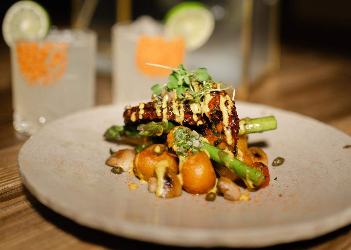 A close-up photograph of “Octopus on the Grill,” a Peruvian fusion dish. Grilled octopus tentacles, drizzled with a yellow sauce, are served on a bed of roasted potatoes and asparagus spears, and garnished with sprouts and capers. Two out-of-focus cocktails with lime wedges and orange-colored garnishes are visible in the background.