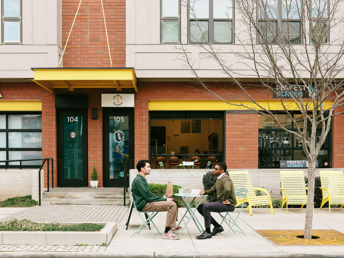 Two men sit at a small mint green bistro table on the sidewalk in front of a modern brick building featuring yellow accents. The storefront is labeled “Wheatberry Bakeshop” above a black door numbered 105, with large windows showing the interior seating area. A leafless tree and several yellow lounge chairs line the walkway, creating a calm, urban atmosphere.