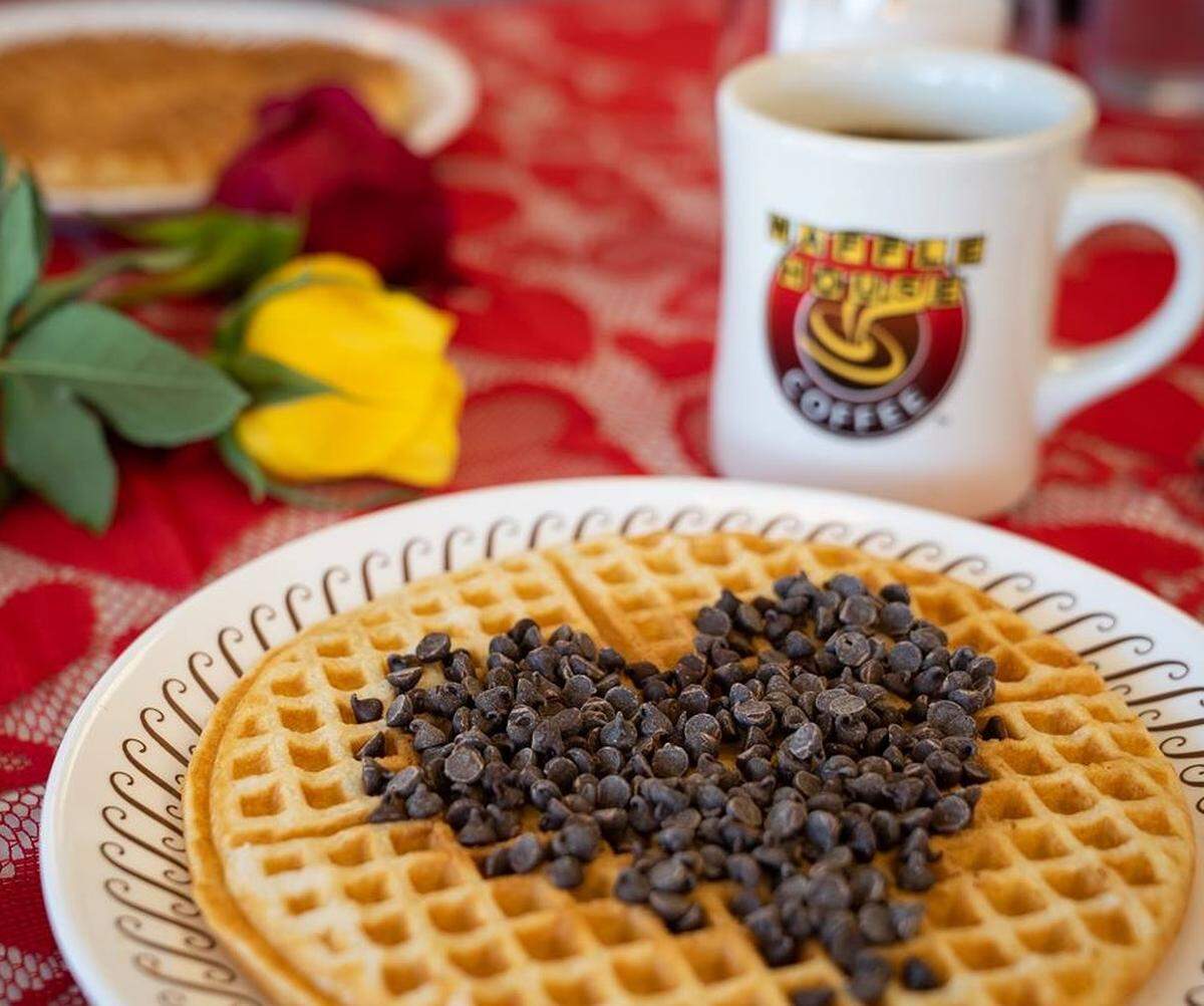 A golden-brown waffle topped with chocolate chips arranged in the shape of a heart sits on a patterned white plate. In the background, a Waffle House coffee mug and colorful roses rest on a red lace tablecloth.