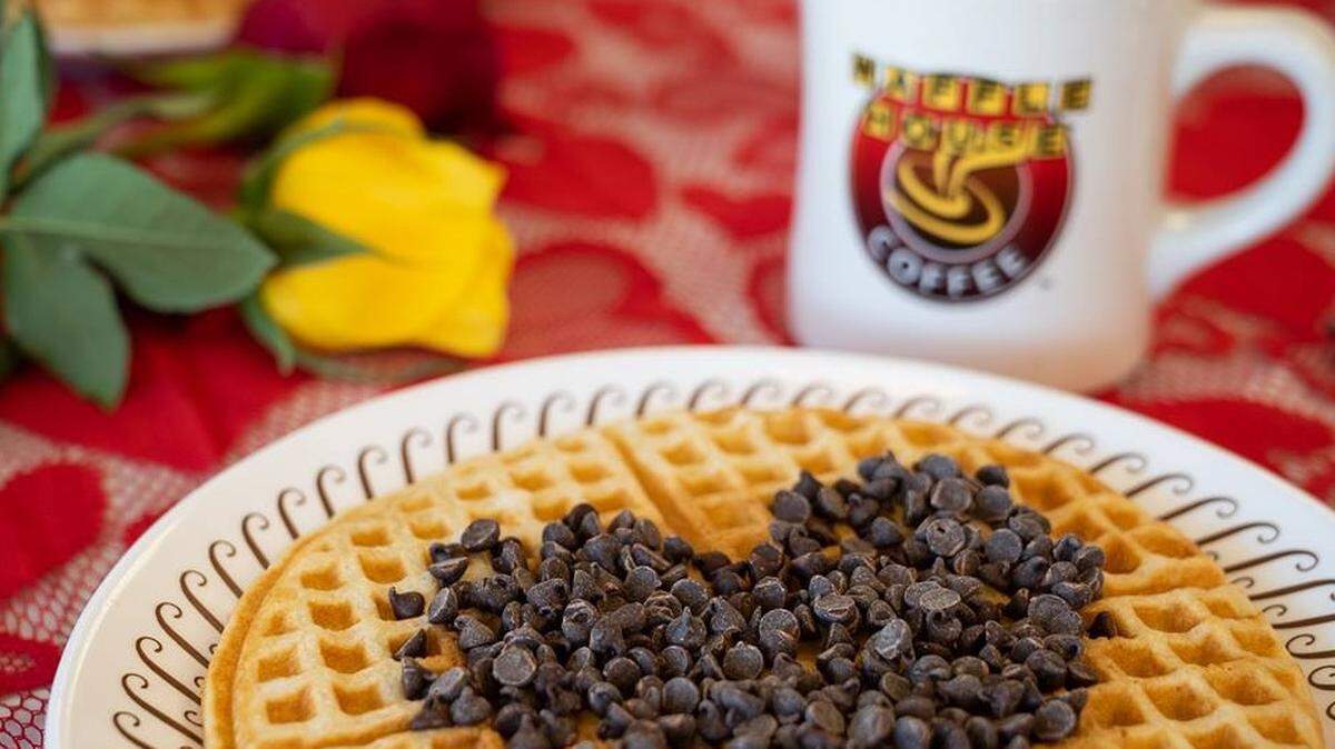 A golden-brown waffle topped with chocolate chips arranged in the shape of a heart sits on a patterned white plate. In the background, a Waffle House coffee mug and colorful roses rest on a red lace tablecloth.