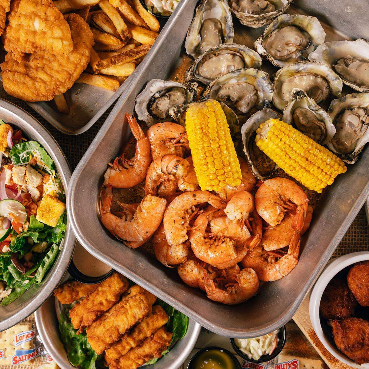 A top-down view of a coastal feast featuring a metal tray filled with steamed shrimp, oysters on the half shell, and corn on the cob. Surrounding the main tray are side dishes including fried fish, french fries, a fresh garden salad, and hushpuppies.