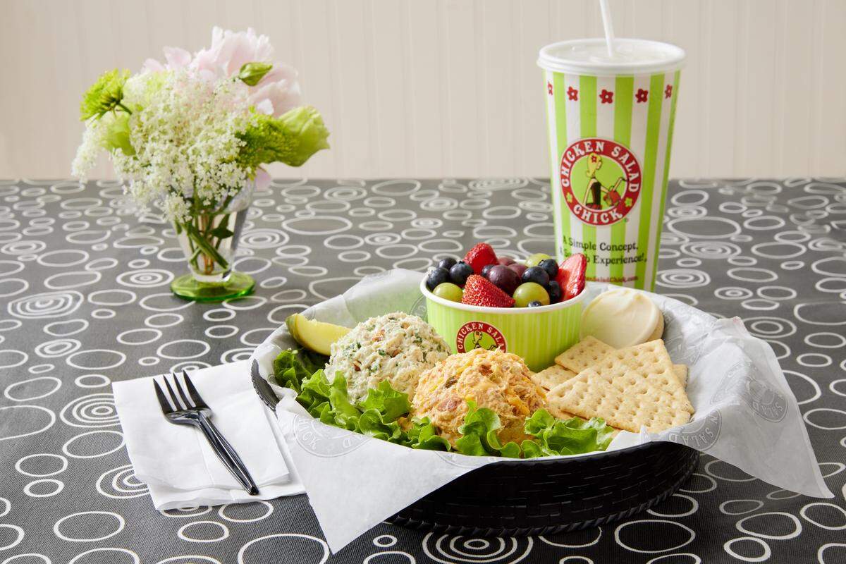 A black wicker basket lined with paper holds two scoops of chicken salad on a bed of lettuce, served with crackers, a pickle, and a cup of fresh fruit. A large green-and-white striped cup and a small vase of flowers sit on the patterned tablecloth in the background.