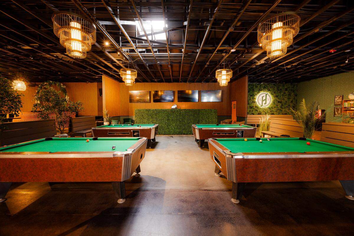 Four green-felted billiard tables are spaced across a large, open entertainment room with polished concrete flooring. The ceiling features exposed black piping and is illuminated by large, tiered woven pendant lights. The back walls are decorated with wood paneling, flat-screen monitors, a privacy hedge, and a green turf accent wall featuring a circular logo.