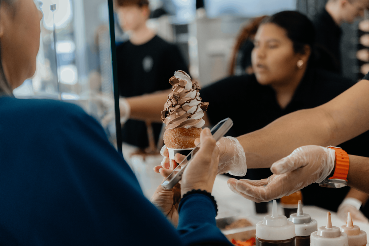 A close-up shot of a customer paying with a smartphone as a worker hands over a swirl of soft-serve ice cream topped with chocolate drizzle and brownie bits, served in a cinnamon-sugar chimney cake cone. The background shows a bustling ice cream shop with other staff members blurred out.