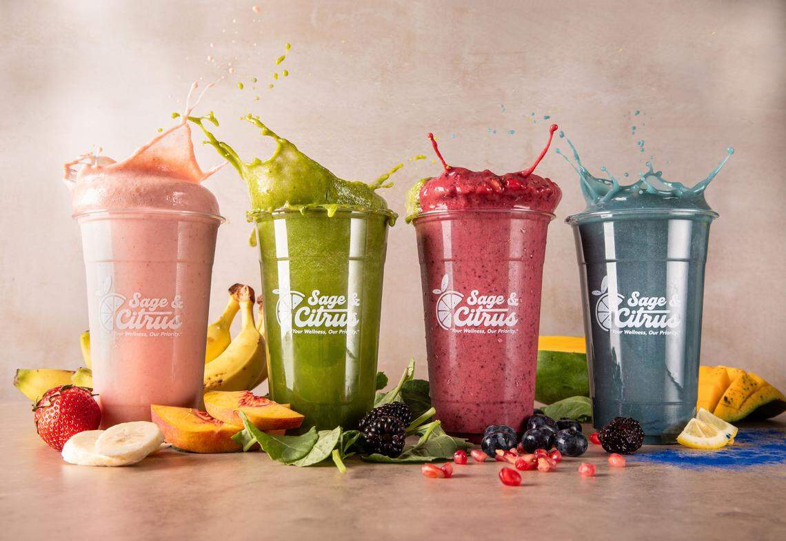 Four vibrant smoothies are lined up in clear cups with the “Sage & Citrus” logo, each color splashing out of the top. The colorful drinks are surrounded by fresh fruit, including strawberries, bananas, peaches, blackberries, and blueberries.