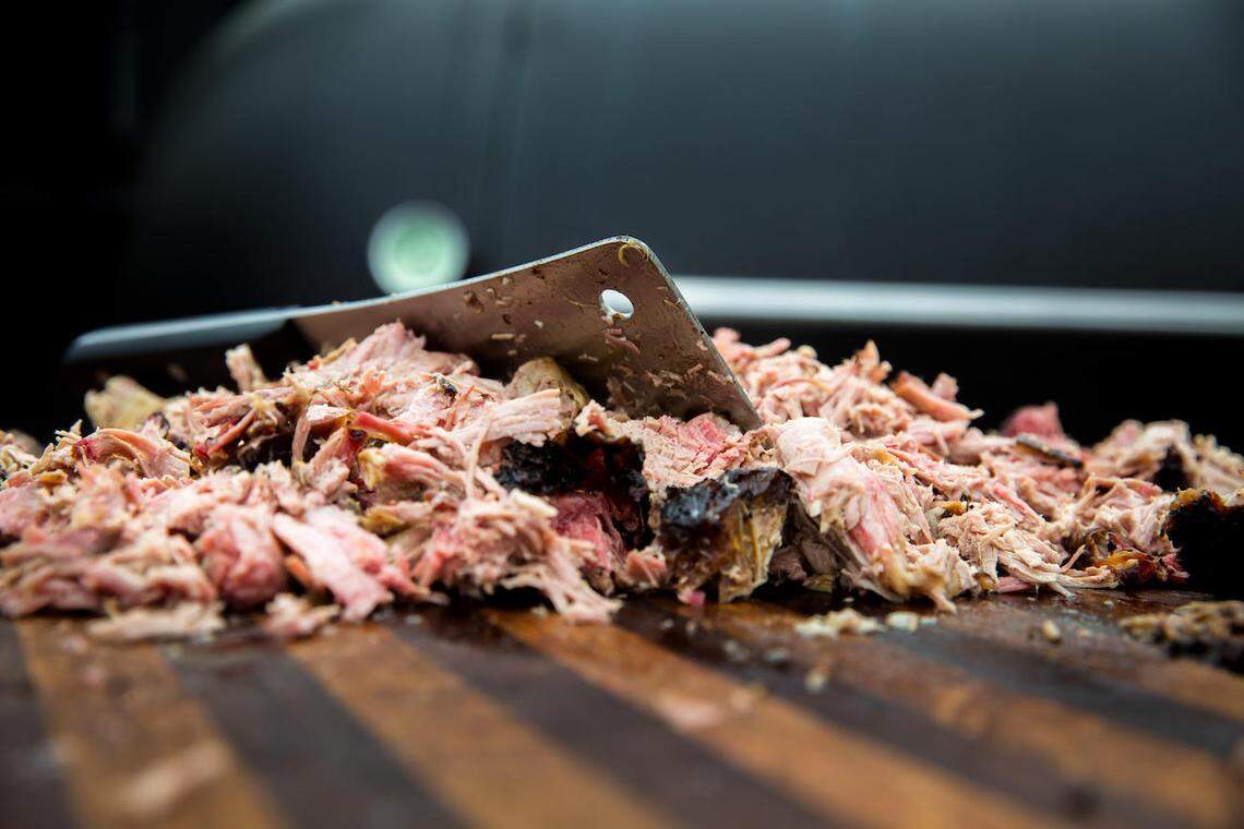 Close-up, low-angle shot of a pile of freshly chopped or pulled smoked pork on a wooden cutting board, with a large, sharp cleaver resting on top of the meat.