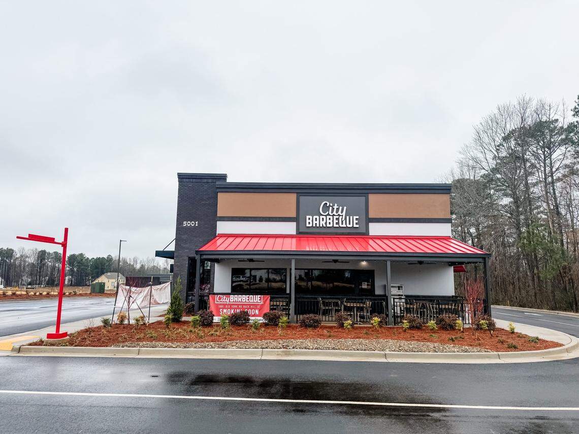 A modern City Barbeque restaurant building features a distinctive bright red metal awning and a “Smokin’ Soon” banner on its outdoor patio. The storefront is situated on a damp asphalt lot under an overcast sky, surrounded by fresh landscaping and pine trees.