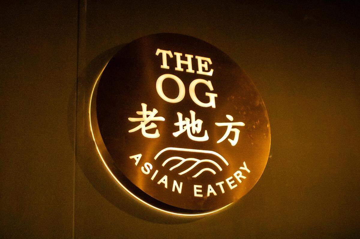 A backlit circular gold sign with the words “THE OG” in English, “老地方” in Chinese characters, and “ASIAN EATERY” underneath with a wavy line graphic. The sign is mounted on a dark wall.