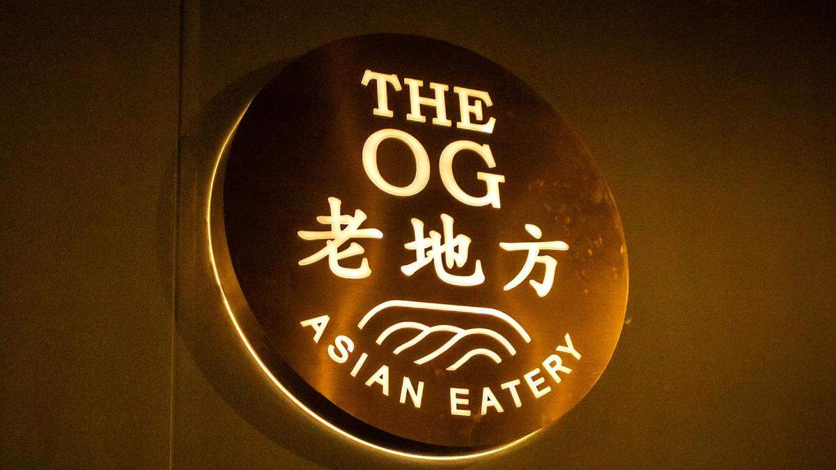 A backlit circular gold sign with the words “THE OG” in English, “老地方” in Chinese characters, and “ASIAN EATERY” underneath with a wavy line graphic. The sign is mounted on a dark wall.