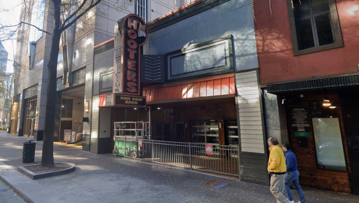 A street-level view shows the entrance of a Hooters restaurant nestled between multi-story urban buildings. A vintage-style vertical neon sign with the iconic owl logo hangs above a black awning that advertises wings, shrimp, and burgers. In the foreground, two pedestrians walk along a brick sidewalk past a metal railing and a patio area with glass garage-style doors.