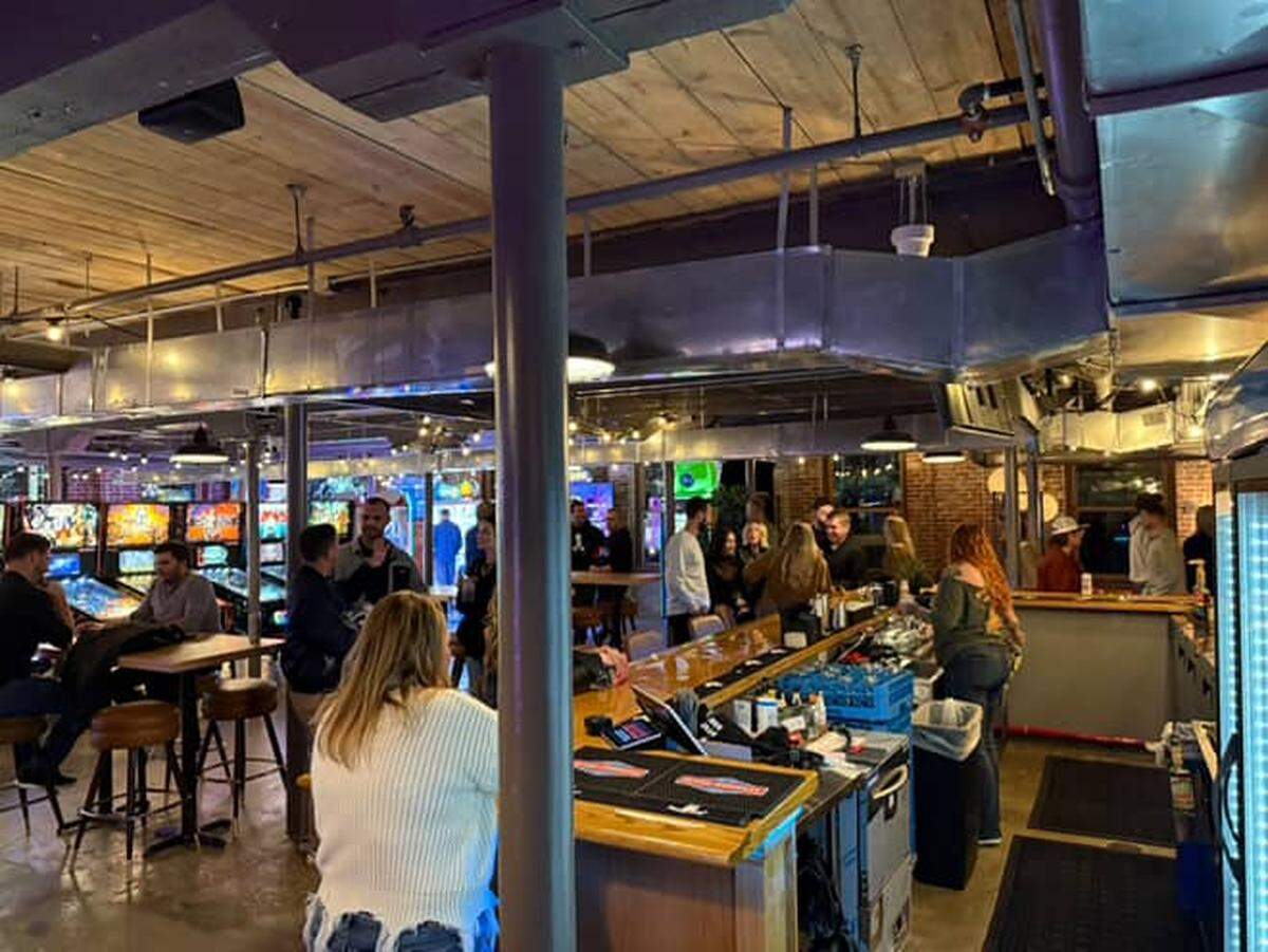 A wide interior shot shows a lively arcade bar with a crowd of people socializing near a central bar and high-top tables. The space features an industrial aesthetic with exposed ductwork, a wood-plank ceiling, metal support beams, and pinball machines visible in the background.