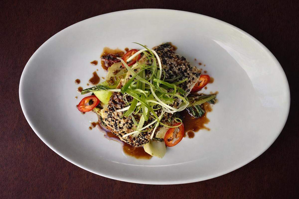 An oval white plate presents a sesame-crusted fish fillet drizzled with dark glaze and garnished with curly green onions and red chili slices. The dish is neatly plated on a dark wood surface, emphasizing a fresh, gourmet presentation.