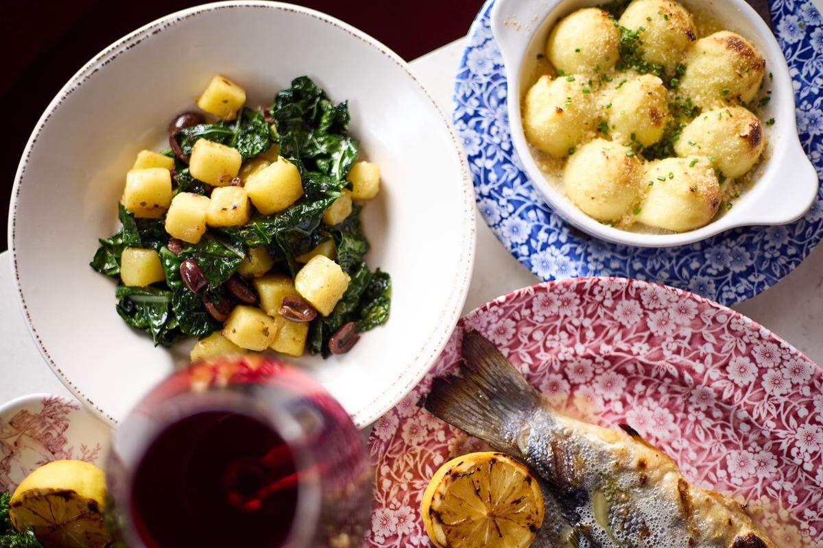 An overhead, close-up shot features three distinct Italian dishes served on patterned ceramic plates. To the left, a bowl holds golden potato gnocchi tossed with dark leafy kale and olives, while a smaller blue floral dish contains toasted, cheesy ravioli. A whole grilled fish with charred lemon sits on a red floral platter in the lower right, partially obscured by a glass of red wine in the foreground.