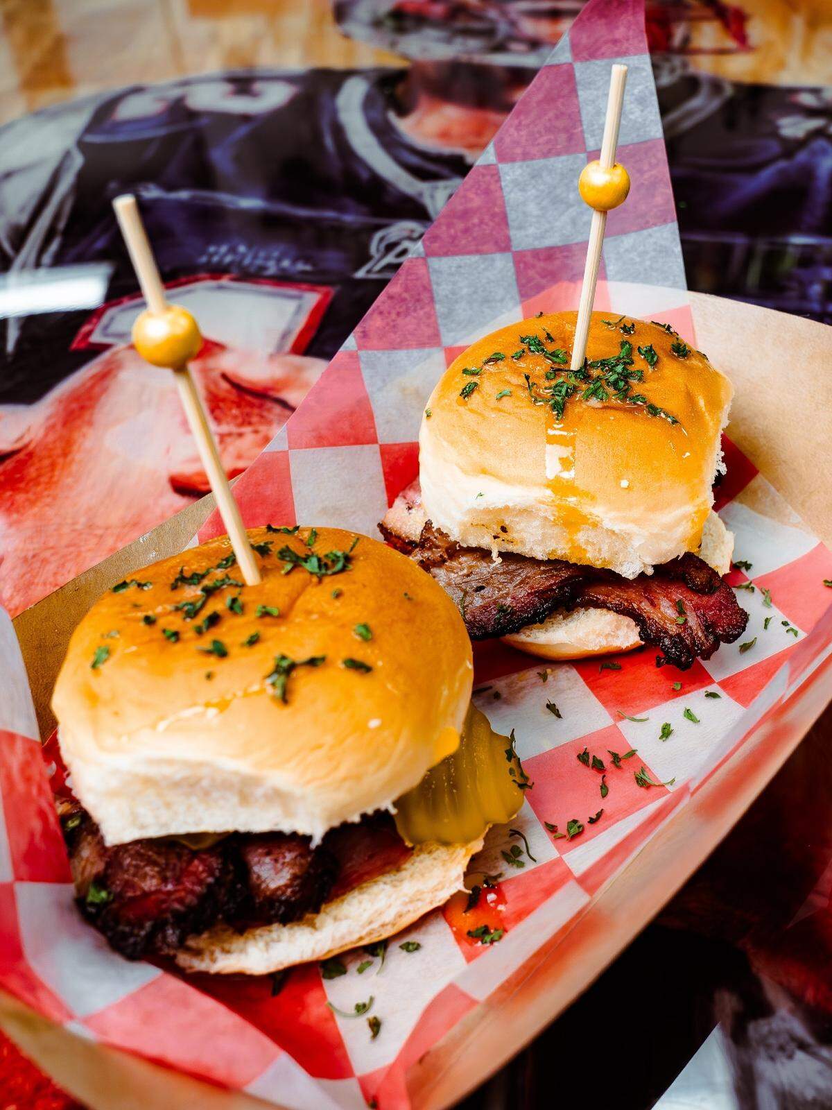 Two brisket sliders with pickles and a honey glaze are served in a red-and-white checkered paper liner. Each slider is held in place by a decorative wooden pick with a gold bead on top.