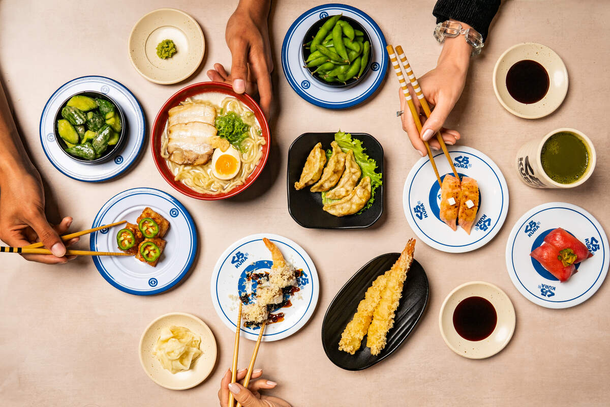 A top-down “flat lay” view captures a large spread of Japanese dishes being shared by three people whose hands and chopsticks are visible. The table is filled with a variety of plates, including a steaming bowl of pork ramen, crispy gyoza, shrimp tempura, and several types of nigiri and rolls. Small side bowls of edamame, ginger, wasabi, and soy sauce complete the communal dining scene.