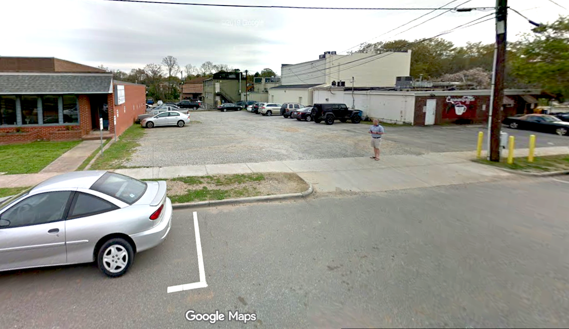 This is a street-level photograph of a gravel parking lot on an overcast day in a small downtown area, with several cars parked in the background. A man in a blue shirt and khaki shorts stands near the center of the lot, which is bordered by a sidewalk, asphalt street, and older commercial buildings.