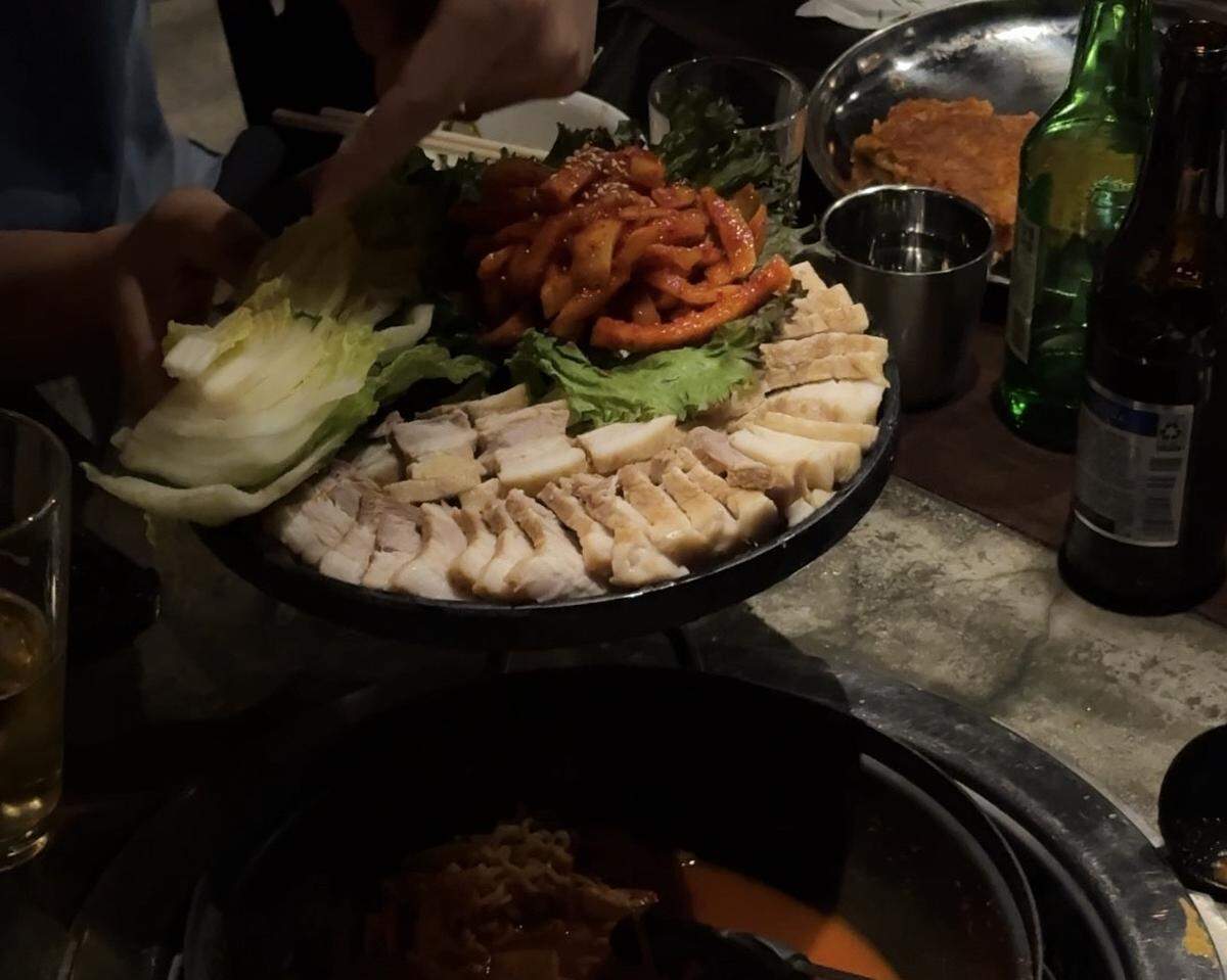 A large platter of sliced boiled pork belly, known as bossam, is served with spicy radish salad and fresh cabbage leaves for wrapping. The table also holds a pot of spicy stew and various alcoholic beverages, capturing a communal dining experience.
