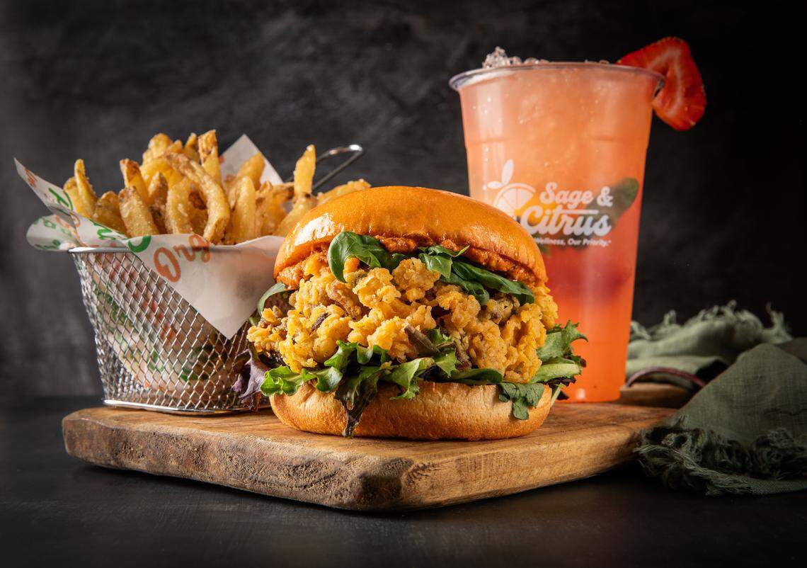 A close-up shot of a combo meal on a wooden cutting board. The meal features a sandwich or burger with a savory-looking, crunchy filling, a wire basket of golden-brown fries, and a pink beverage garnished with a strawberry slice.