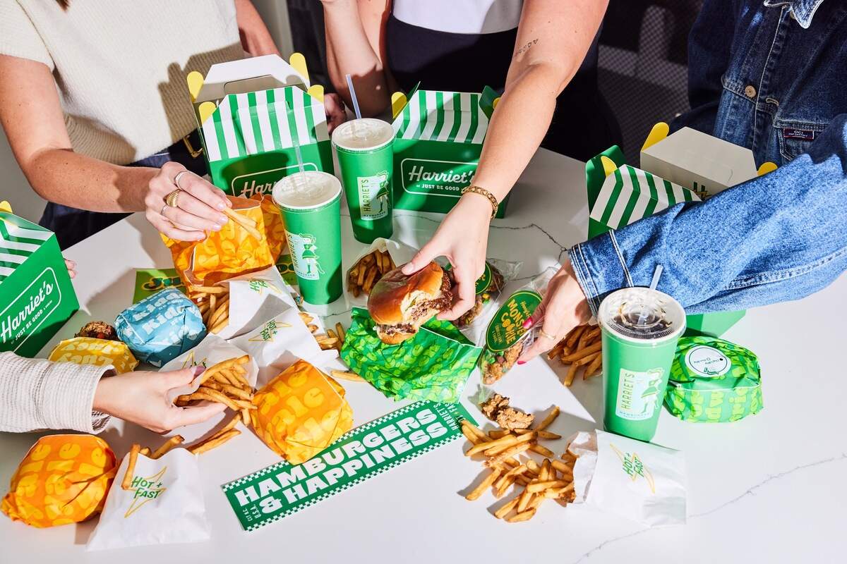 A high-angle shot shows a white table filled with a spread of fast food from Harriet’s Hamburgers, surrounded by four people sharing the meal. Green and white striped takeout boxes, bright green cups, and various wrapped items are scattered across the table next to loose french fries. One person holds a half-eaten burger over a green wrapper, while others reach for fries and a packaged cookie.