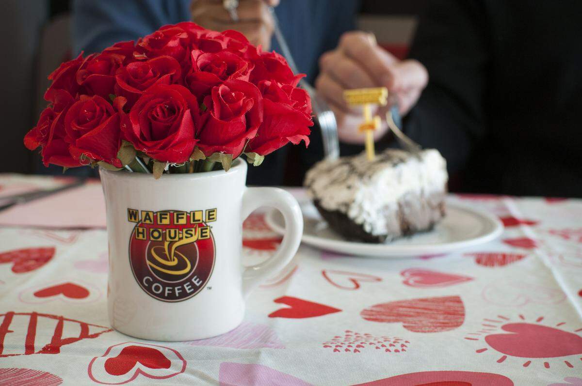 A white Waffle House coffee mug holds a bouquet of vibrant red roses on a table covered in a heart-patterned cloth. In the blurred background, a person prepares to eat a slice of pie topped with whipped cream.