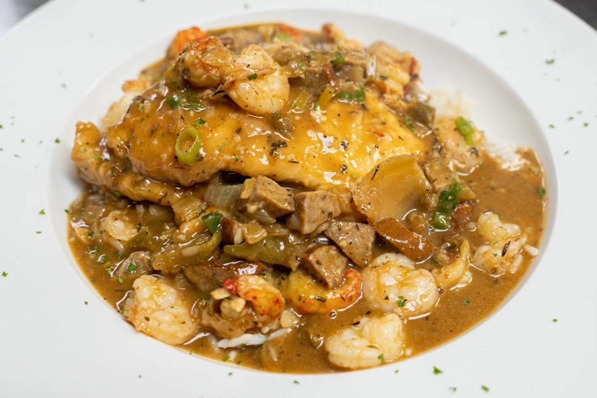 A close-up of a Cajun-style dish featuring a piece of blackened or grilled fish topped with a rich, brown etouffee sauce. The sauce is filled with plump shrimp, chunks of crawfish or sausage, and sautéed green bell peppers and onions. The dish is served over a bed of white rice in a shallow white bowl and garnished with a sprinkle of fresh parsley.
