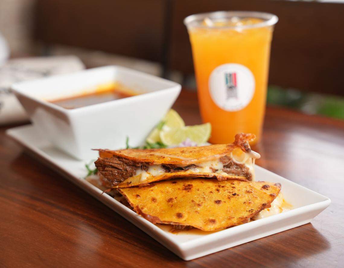 Two folded, cheese-covered Birria tacos with crispy orange shells and meat filling are presented on a long white rectangular plate, garnished with lime wedges and cilantro. Beside the tacos, a white bowl holds a portion of rich red consommé, and a tall plastic cup of bright orange juice stands in the background.