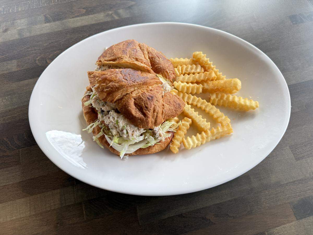 A white plate holding a croissant sandwich filled with tuna or chicken salad and lettuce, served next to a handful of crinkle-cut french fries.