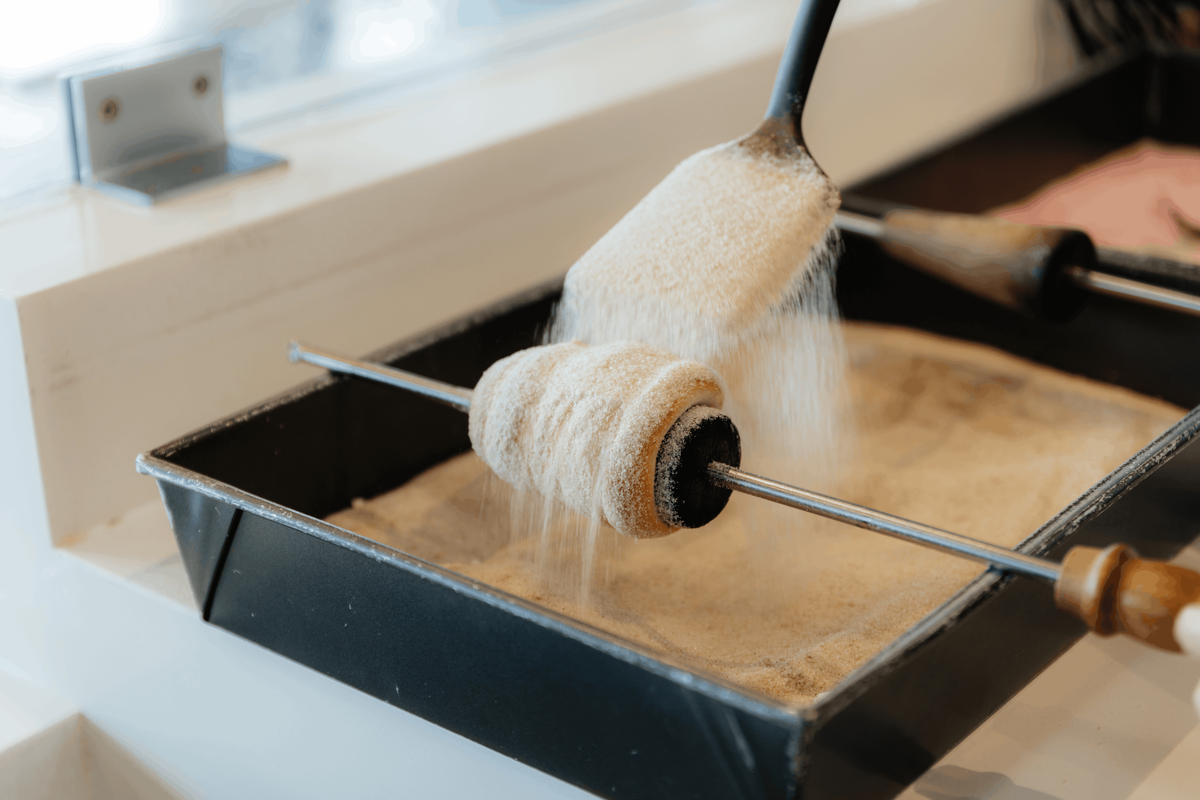 A close-up shows a fresh chimney cake pastry rotating on a metal spit over a metal tray as cinnamon sugar is scooped and poured generously over it. The motion captures the sugar cascading over the golden-brown dough in a busy bakery setting.