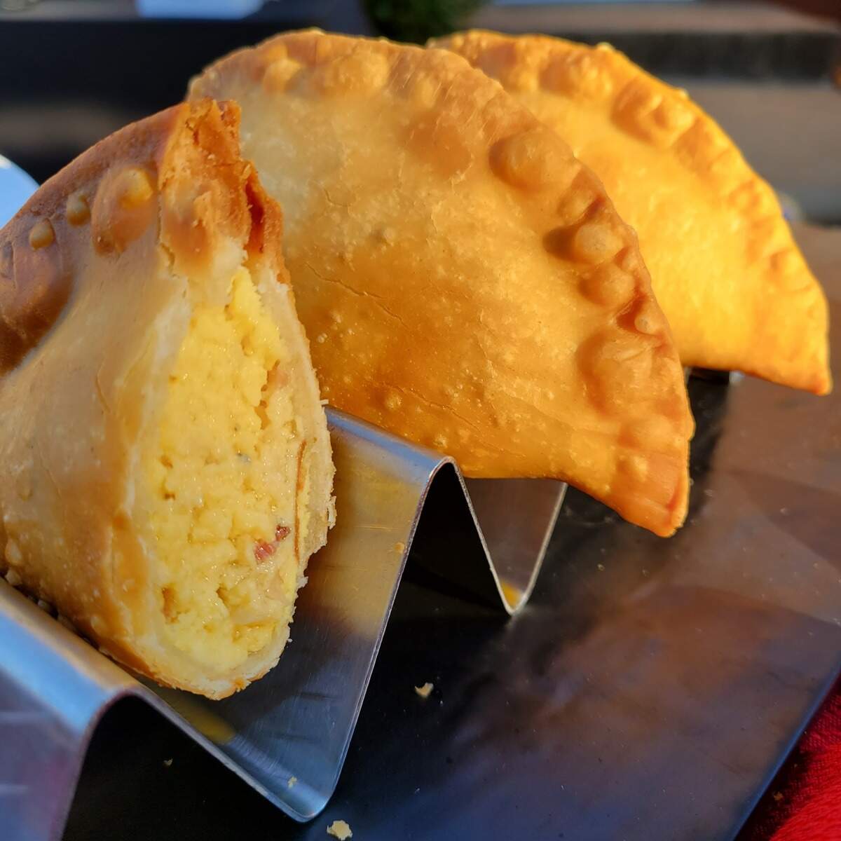 Two golden-brown empanadas rest in a metal holder, with one cut open to reveal a creamy, pale-yellow filling. The close-up highlights the crimped edges and blistered crust.