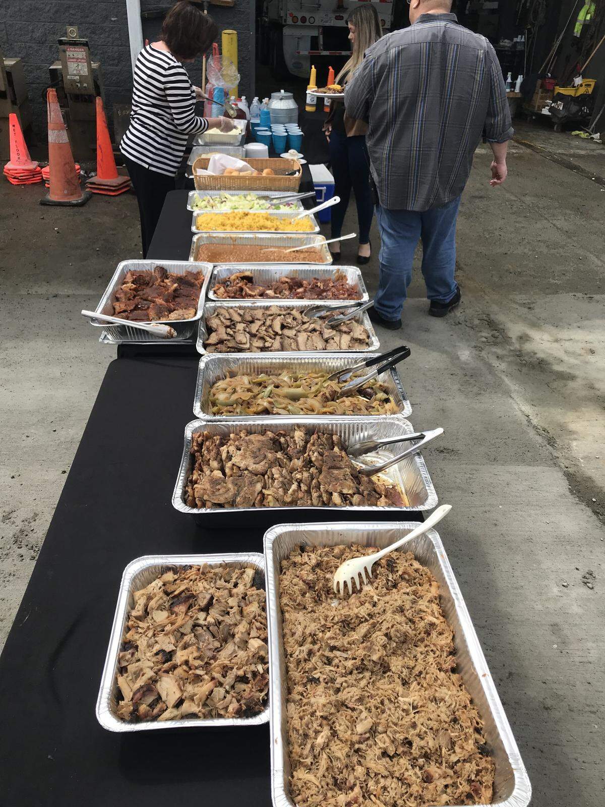 This image captures a buffet line set up, featuring several large aluminum trays filled with different types of barbecued and pulled meats, along with sides like shredded lettuce and cheese. In the background, people are preparing and serving food from the far end of the long black-covered table, suggesting a catered event or gathering.