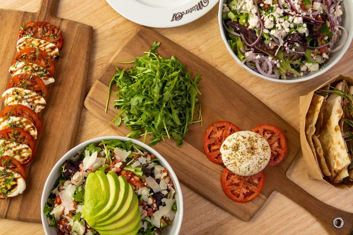 An overhead view shows several wooden boards and bowls filled with caprese skewers, a burrata salad, fresh arugula, and Mediterranean-style salads.