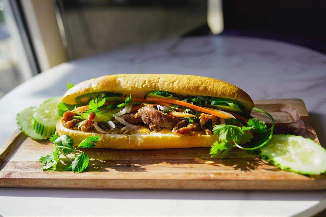 A close-up shot features a vibrant Bánh Mì sandwich on a wooden serving board, packed with grilled meat, pickled carrots, cucumbers, and fresh cilantro. The crusty baguette is perfectly toasted and sits under bright, natural sunlight that highlights the fresh ingredients.