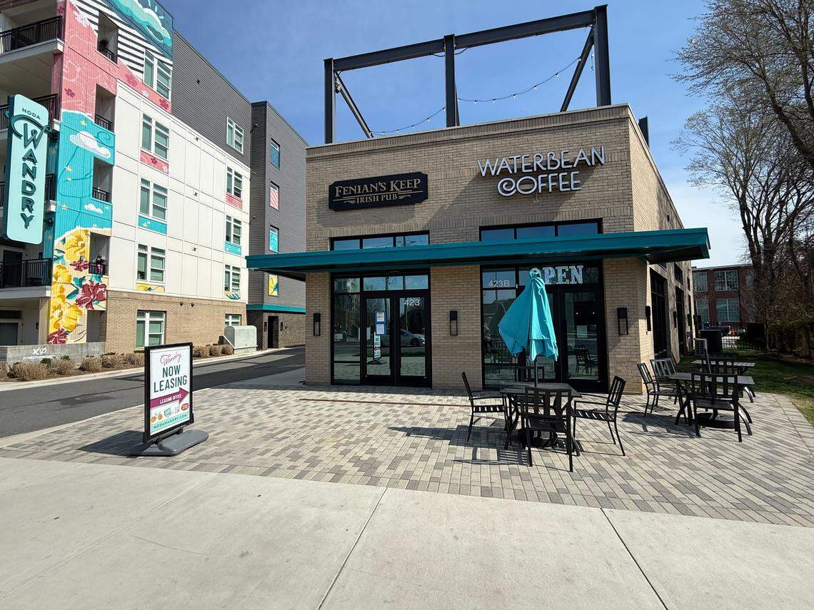 A wide-angle shot shows a tan brick building housing Fenian’s Keep Irish Pub and Waterbean Coffee, situated next to a modern apartment complex with a vibrant floral mural. The foreground features a paved patio with outdoor seating and a “Now Leasing” sign for the Noda Wandry apartments.