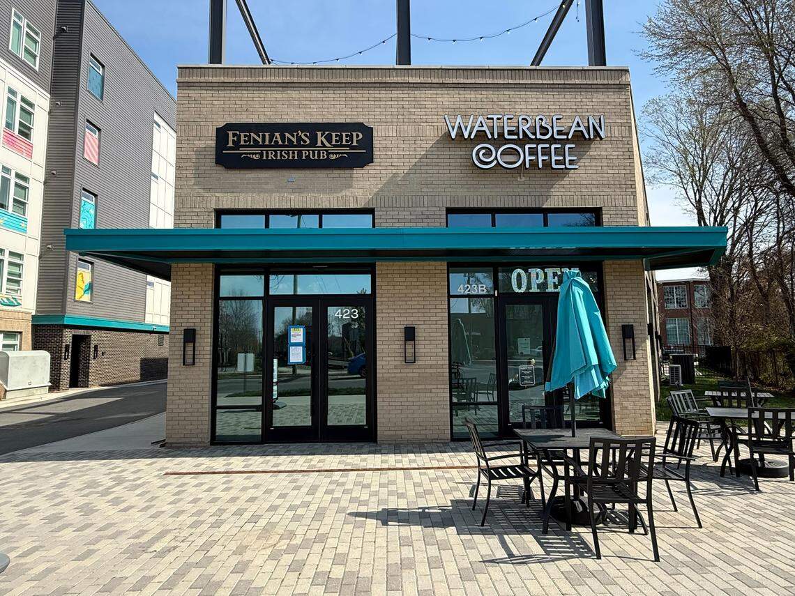 This eye-level medium shot captures the dual storefronts of Fenian’s Keep Irish Pub and Waterbean Coffee under a clear blue sky. A teal awning stretches across the front of the building, shading a patio area with black metal tables and a closed teal umbrella.