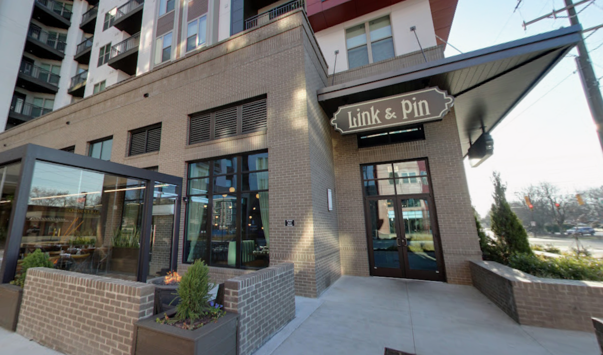 Exterior view of Link & Pin restaurant, located in the ground floor of a modern, multi-story brick apartment building. The entrance is marked by a black awning with the “Link & Pin” logo. The building features large windows and an outdoor seating area enclosed by glass panels and a low brick wall.