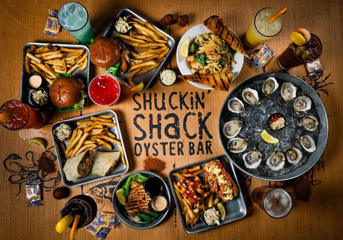A large wooden table features the “Shuckin’ Shack Oyster Bar” logo surrounded by an extensive variety of menu items. The spread includes raw oysters, burgers, shrimp pasta, fish tacos, and several colorful cocktails and beers.