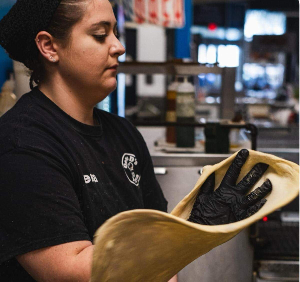 A woman in a black t-shirt and gloves carefully stretches out a circular piece of pizza dough in a professional kitchen. She focuses intently on the dough, which is draped over her hands as she prepares it for toppings.