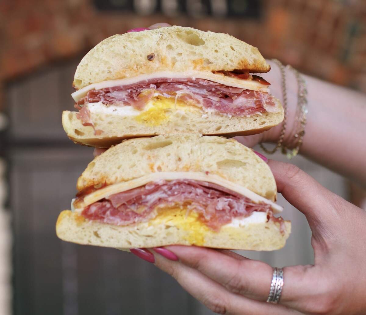 A close-up, front-facing shot shows a person holding the two halves of a breakfast sandwich stacked on top of each other. The sandwich is made on a fluffy roll and layered with sliced cheese, ribbons of Italian cured meat, and a fried egg with a bright yellow yolk. The background is softly blurred, focusing attention on the textures of the food.