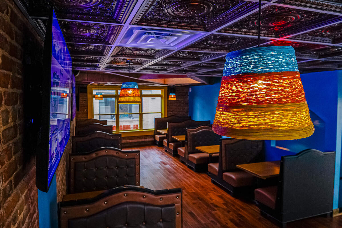 A moody restaurant interior showcases a long row of dark brown, tufted leather dining booths lining a polished hardwood floor. The ceiling is covered in ornate, metallic pressed tin tiles, illuminated by a mix of blue ambient lighting and colorful, woven cone-shaped pendant lamps. In the background, a large yellow-framed window looks out onto the street, while a massive flat-screen television mounts to the brick wall on the left.