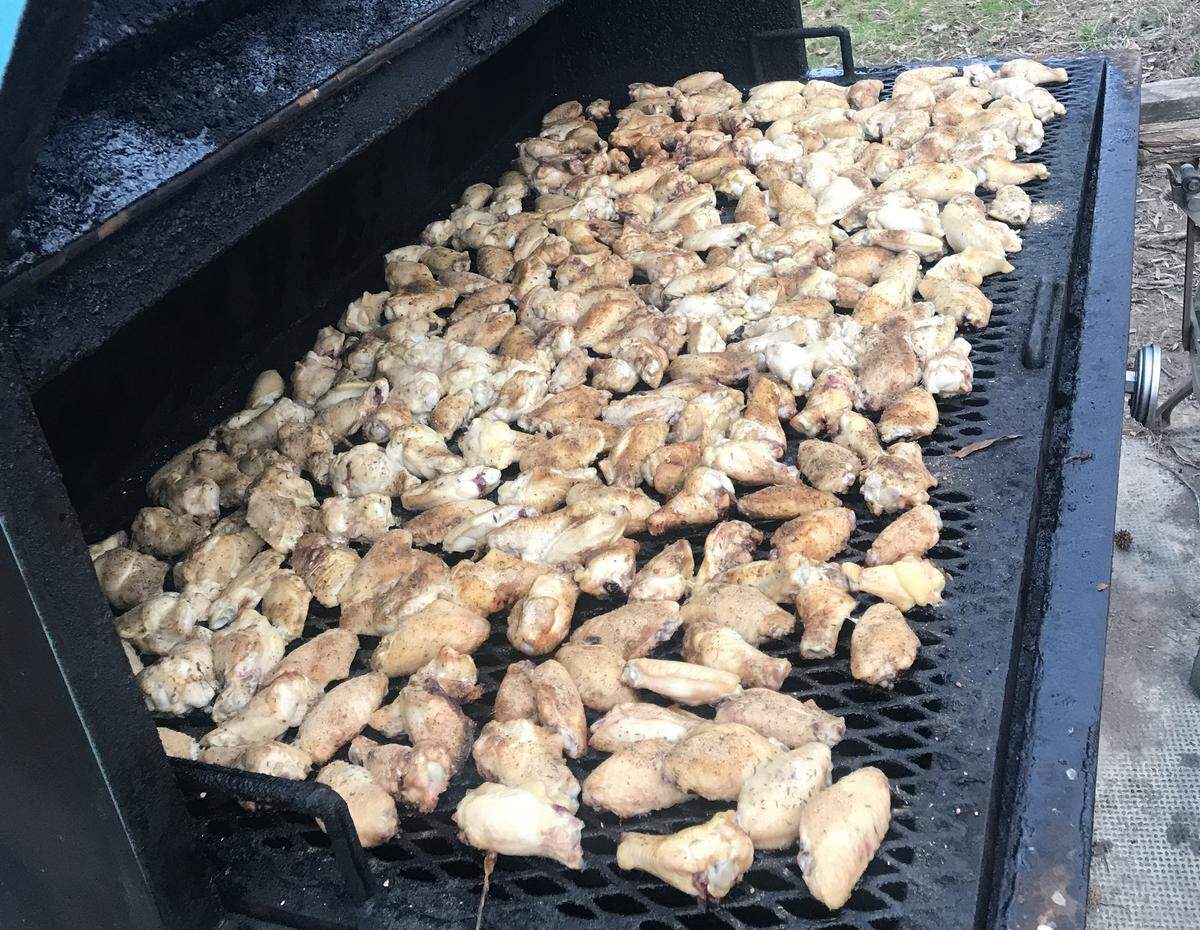 This photo shows a massive outdoor smoker, with the lid propped open, filled with hundreds of uncooked, seasoned chicken wings spread across the grill grates. The dark, heavily used smoker and the quantity of meat suggest this is a professional setup for large-volume cooking.