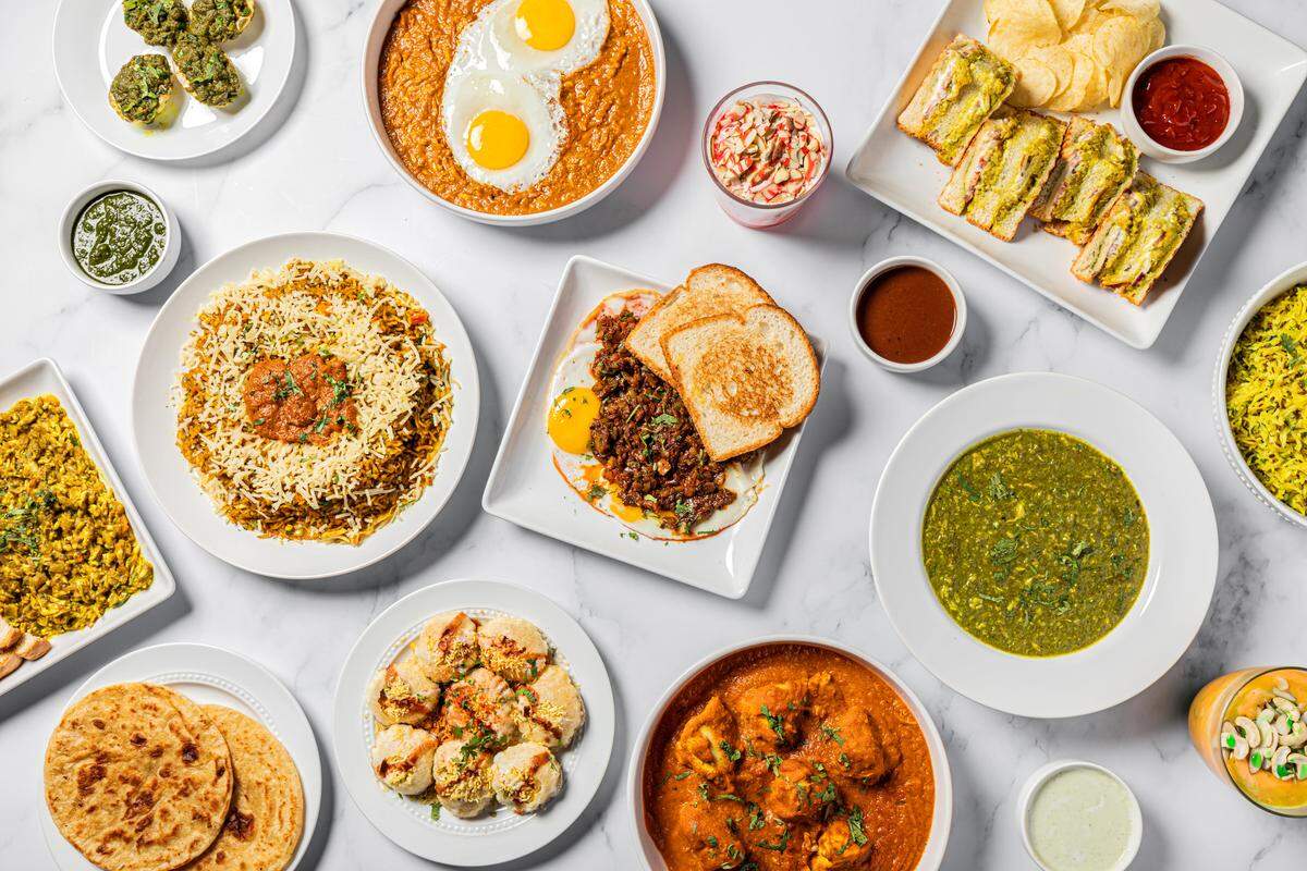 An overhead flat lay of a large assortment of Indian food arranged on a white marble table. Dishes include rice, various curries, chicken and egg preparations, flatbreads, savory snacks, and beverages.