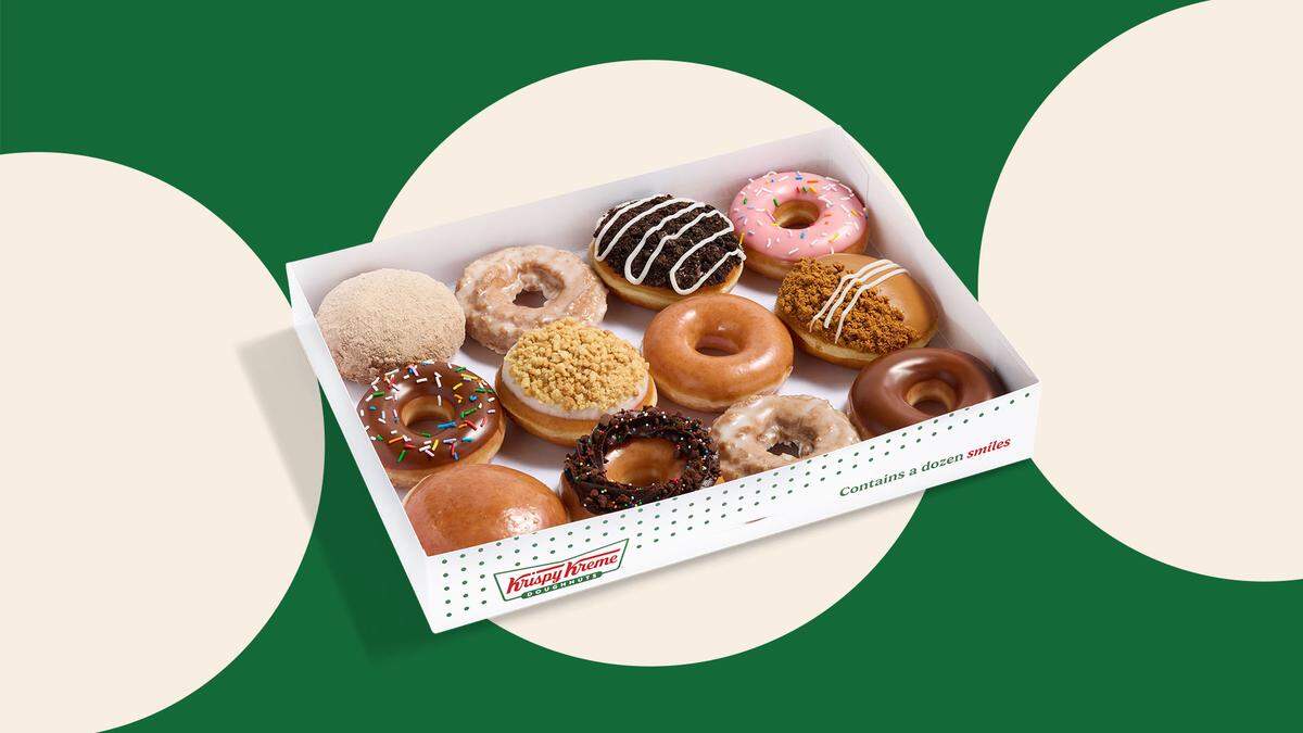 This image shows a white Krispy Kreme dozen box opened up, containing a variety of twelve different doughnuts with various glazes, toppings, and fillings. The box is placed against a background of solid green and large cream-colored circular shapes, highlighting the bright colors and textures of the pastries.