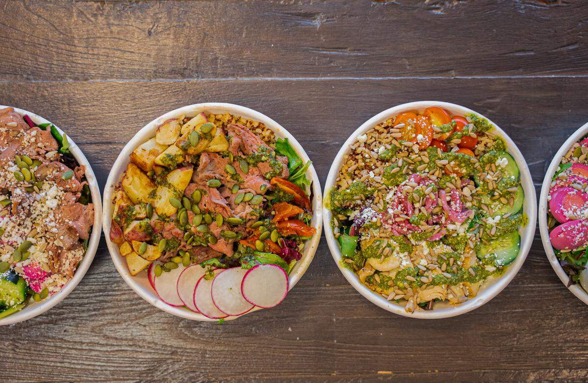 Four overhead bowls are lined up on a dark wooden surface, showcasing variety in their ingredients. The bowls are packed with colorful savory items including sliced radishes, roasted potatoes, leafy greens, and various seeds or grains.