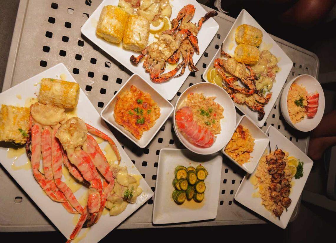 A metal table is covered with an abundant seafood feast featuring multiple plates of grilled crab, lobster tails, and snow crab legs. Side dishes including corn on the cob, mac and cheese, and sliced zucchini are tucked between the main platters. The overhead view showcases a variety of textures and golden-brown finishes, creating a lavish spread ready for a group.