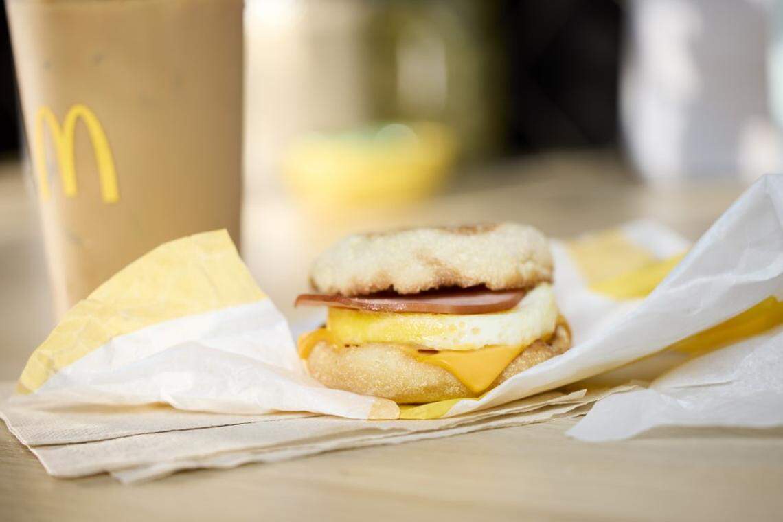 A McDonald’s Egg McMuffin rests on its yellow and white wrapper atop a stack of brown napkins, showcasing its layers of toasted English muffin, Canadian bacon, a round cracked egg, and melted American cheese. In the soft-focus background, a large McCafé iced coffee sits on a wooden table, completing the classic breakfast scene.
