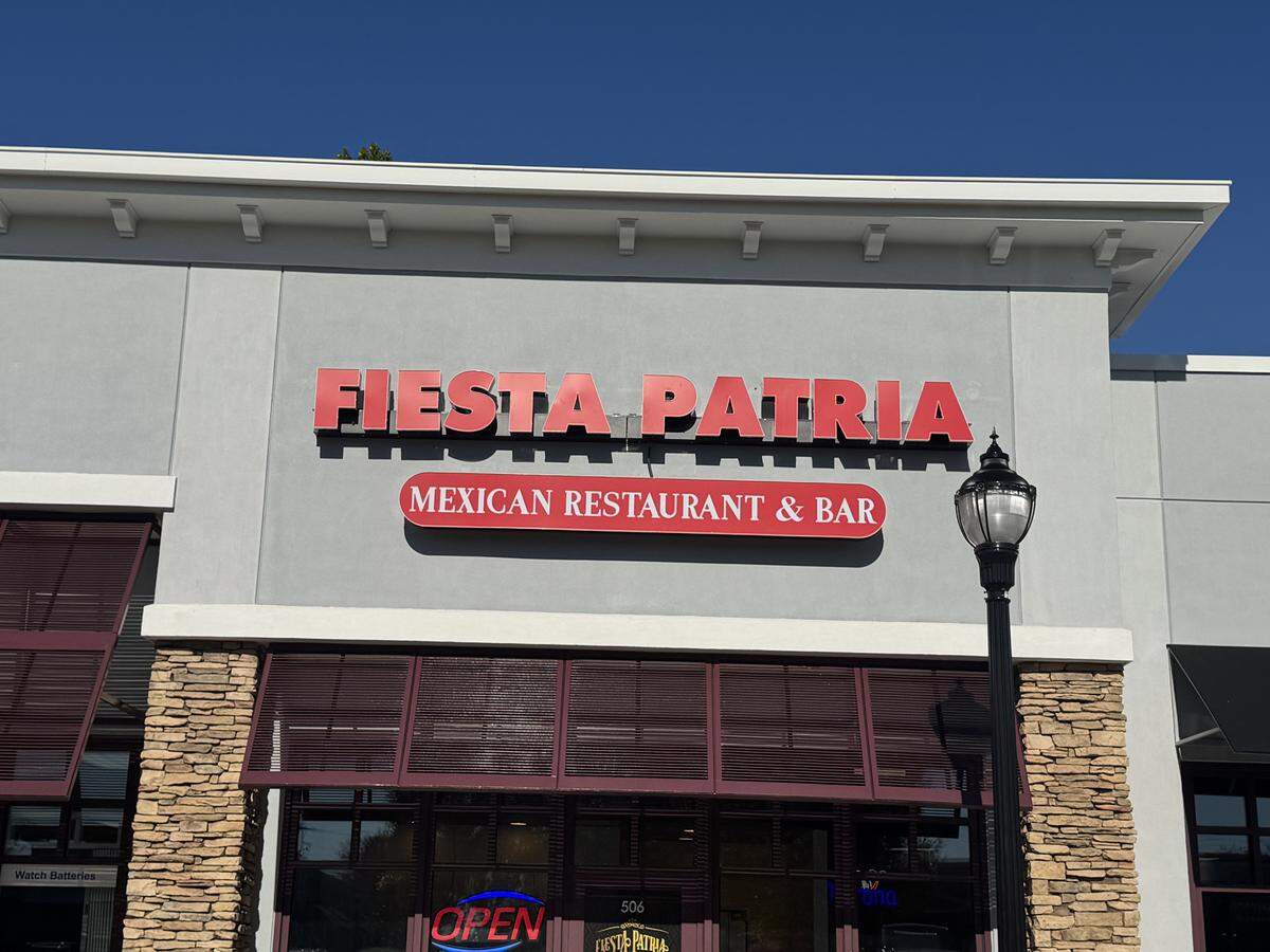 The exterior of the restaurant features a gray facade with a section of stacked stone below, all under a clear blue sky. Red dimensional letters spell out “FIESTA PATRIA” above a red oval sign that reads “MEXICAN RESTAURANT & BAR.”